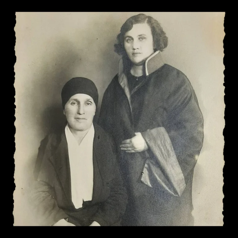 Studio portrait of a seated elderly woman with her head covered and, standing beside her, a young woman in dark, wide-collared clothes