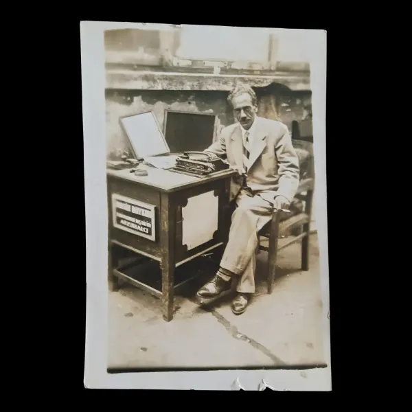 Hombre con traje de color claro sentado en una silla junto a su mesa con máquina de escribir y el letrero 'Arzuhalci' (memorialista)