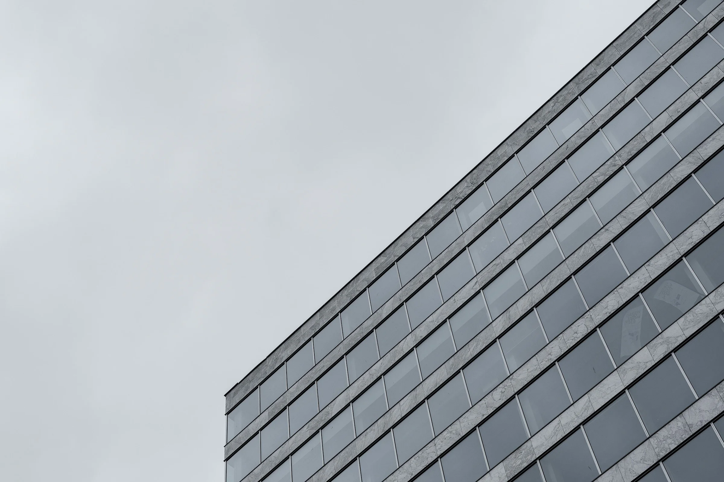 Marble-clad office building facade with glass windows, rising diagonally toward a cloudy sky